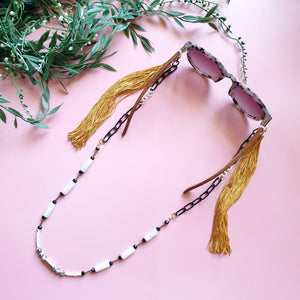 A stylish and multifunctional accessory that can be worn as a sunglasses chain, necklace, or lanyard. Featuring a mix of black and white chain links, delicate beads, and long flowing gold fringe for a bold and playful touch.