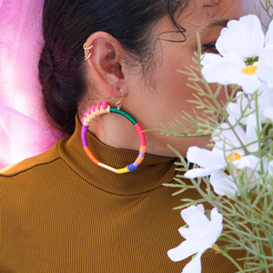 Model is wearing Brave hoop earrings with pink pompom wrapped in peach and yellow thread, accented with gold plate, paired with white mesh fabric around her head and neck, highlighting the vibrant colours of the earrings.