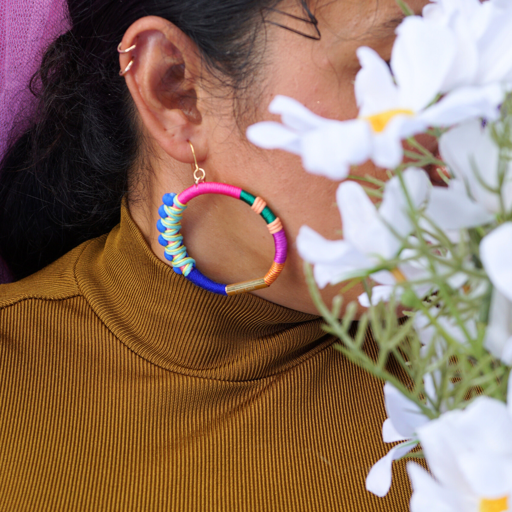 Model is wearing colourful hoop earrings wrapped in vibrant thread with gold-tone details and blue pom-poms