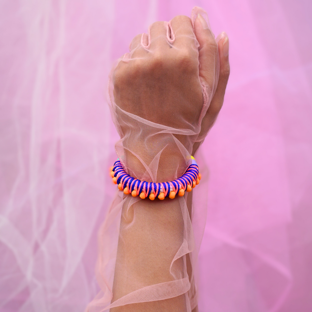 A close-up of a hand adorned with vibrant, handmade bracelets wrapped in colorful thread, featuring tassels and beads. The hand is elegantly posed against a soft pink sheer fabric background, creating a bold and playful aesthetic. Perfect for a bohemian or festival-inspired jewelry collection.