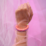 A close-up of a hand adorned with vibrant, handmade bracelets wrapped in colorful thread, featuring tassels and beads. The hand is elegantly posed against a soft pink sheer fabric background, creating a bold and playful aesthetic. Perfect for a bohemian or festival-inspired jewelry collection.