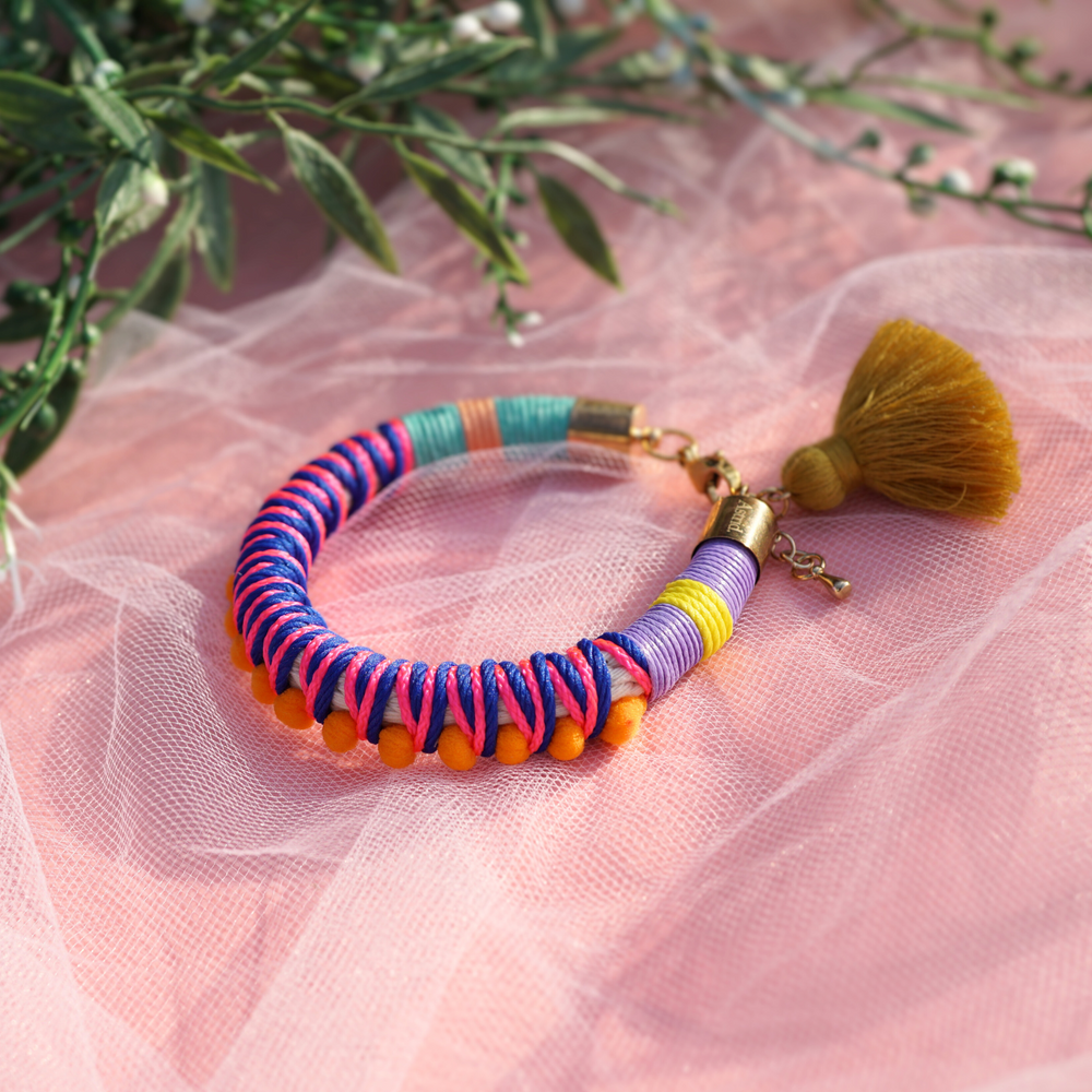 A close-up of a hand adorned with vibrant, handmade bracelets wrapped in colorful thread, featuring tassels and beads. The hand is elegantly posed against a soft pink sheer fabric background, creating a bold and playful aesthetic. Perfect for a bohemian or festival-inspired jewelry collection.