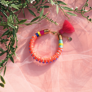 A vibrant handmade bracelet featuring lilac pom-poms, multicolored thread wrapping, and a pink tassel charm, displayed on soft pink tulle with greenery in the background. This boho-chic statement piece adds a playful and colorful touch to any outfit, perfect for festival and summer styling