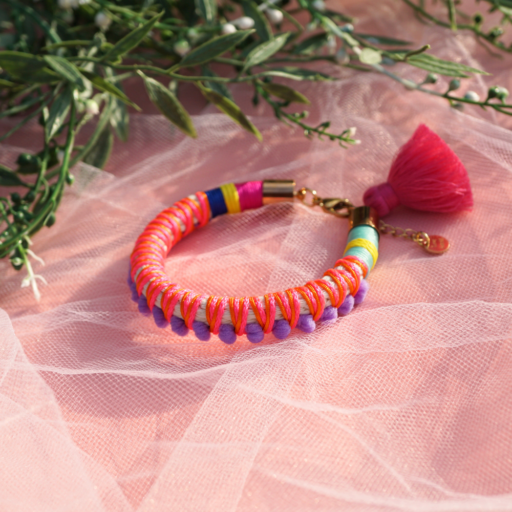 A vibrant handmade bracelet featuring lilac pom-poms, multicolored thread wrapping, and a pink tassel charm, displayed on soft pink tulle with greenery in the background. This boho-chic statement piece adds a playful and colorful touch to any outfit, perfect for festival and summer styling