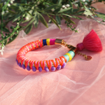 A close-up of a hand adorned with vibrant, handmade bracelets wrapped in colorful thread, featuring tassels and beads. The hand is elegantly posed against a soft pink sheer fabric background, creating a bold and playful aesthetic. Perfect for a bohemian or festival-inspired jewelry collection.