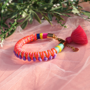 A vibrant handmade bracelet featuring lilac pom-poms, multicolored thread wrapping, and a pink tassel charm, displayed on soft pink tulle with greenery in the background. This boho-chic statement piece adds a playful and colorful touch to any outfit, perfect for festival and summer styling