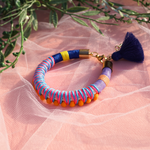 A close-up of a hand adorned with vibrant, handmade bracelets wrapped in colorful thread, featuring tassels and beads. The hand is elegantly posed against a soft pink sheer fabric background, creating a bold and playful aesthetic. Perfect for a bohemian or festival-inspired jewelry collection.