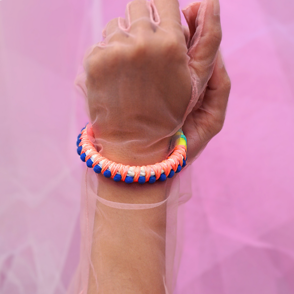 A close-up of a hand wearing an orange and blue pom-pom bracelet, wrapped in colorful thread. The handmade bracelet features intricate woven details, adding a bold and playful touch to any outfit. Perfect for boho, festival, or statement jewellery lovers.