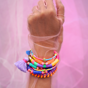 A close-up of a hand adorned with vibrant, handmade bracelets wrapped in colorful thread, featuring tassels and beads. The hand is elegantly posed against a soft pink sheer fabric background, creating a bold and playful aesthetic. Perfect for a bohemian or festival-inspired jewelry collection.