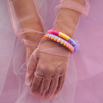 A close-up of a hand adorned with vibrant, handmade bracelets wrapped in colorful thread, featuring tassels and beads. The hand is elegantly posed against a soft pink sheer fabric background, creating a bold and playful aesthetic. Perfect for a bohemian or festival-inspired jewelry collection.