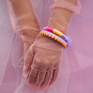 A pair of handmade, colorful pom-pom bracelets stacked on a model's wrist, wrapped in sheer pink gloves. The bracelets feature bright orange, lilac, and multicolored thread details, creating a bold and playful boho-chic statement. Perfect for summer, festivals, and vibrant accessorizing.