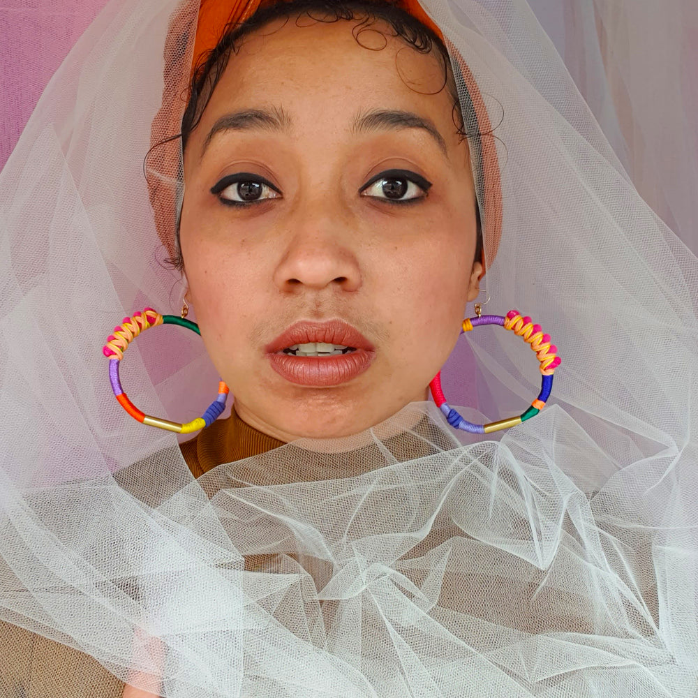 Model is wearing Brave hoop earrings with pink pompom wrapped in peach and yellow thread, accented with gold plate, paired with white mesh fabric around her head and neck, highlighting the vibrant colours of the earrings.