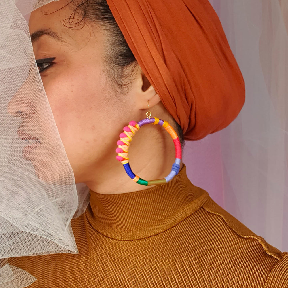 Model showcasing Brave hoop earrings, accentuating their vibrant and colourful design with pink pompom wrapped in peach and yellow thread, and gold plate accents.