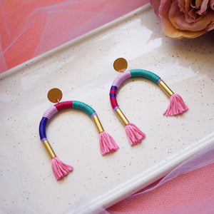 Colourful arch earrings wrapped in lilac, blue, and pink thread with gold tube accents, featuring hand-dyed pink tassels at each end.