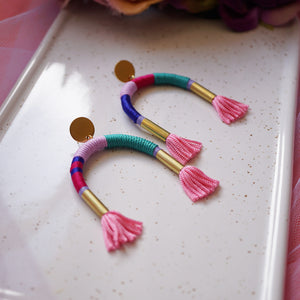 Colourful arch earrings wrapped in lilac, blue, and pink thread with gold tube accents, featuring hand-dyed pink tassels at each end.