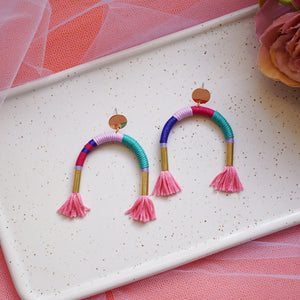 Colourful arch earrings wrapped in lilac, blue, and pink thread with gold tube accents, featuring hand-dyed pink tassels at each end.