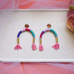 Colourful arch earrings wrapped in lilac, blue, and pink thread with gold tube accents, featuring hand-dyed pink tassels at each end.