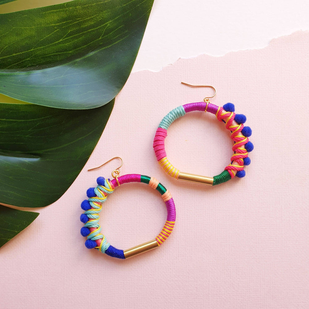 Colourful hoop earrings wrapped in vibrant pink, blue, green, and yellow thread, accented with small blue pom-poms and gold-tone metal details