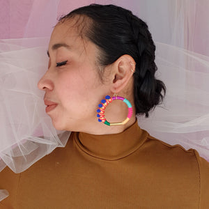 Model is wearing colourful hoop earrings wrapped in vibrant thread with gold-tone details and blue pom-poms