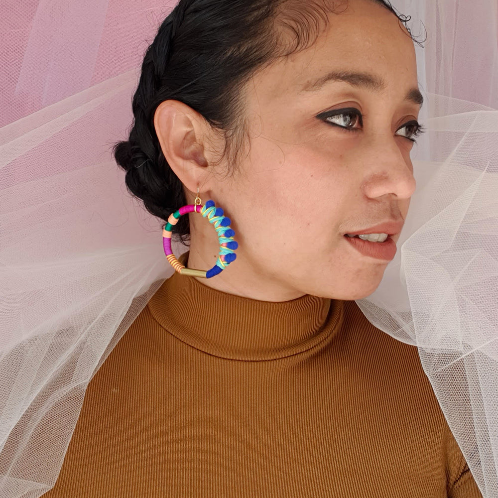 Model is wearing colourful hoop earrings wrapped in vibrant thread with gold-tone details and blue pom-poms