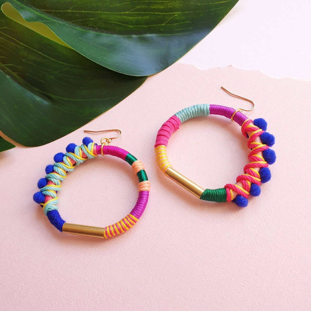 Colourful hoop earrings wrapped in vibrant pink, blue, green, and yellow thread, accented with small blue pom-poms and gold-tone metal details