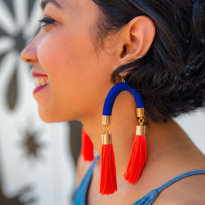 A model wears bold handmade earrings with striking blue arches and vibrant orange tassels, adding a statement touch to her look.