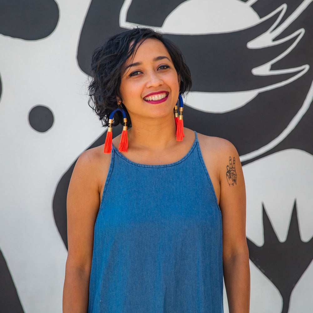 A model wears bold handmade earrings with striking blue arches and vibrant orange tassels, adding a statement touch to her look.