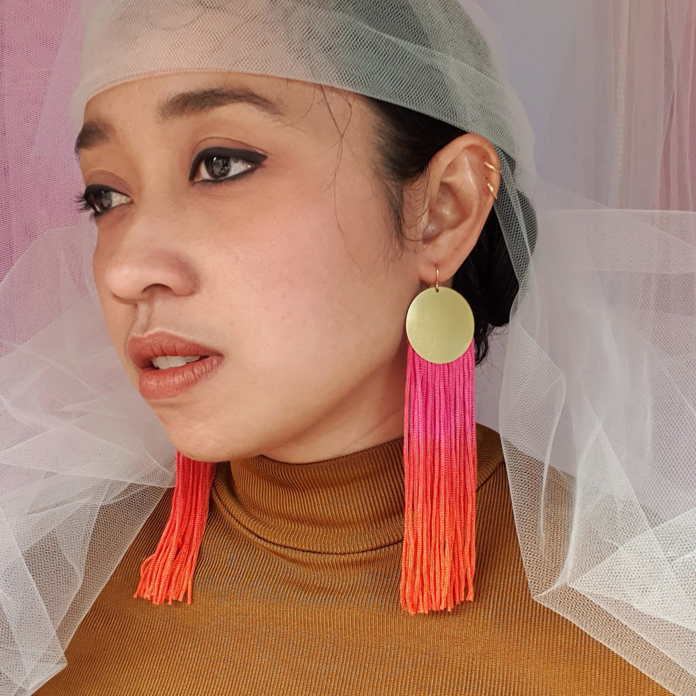 A model wears elegant pink and orange ombre tassel earrings with gold-plated round pendants. The bold, vibrant tassels add a statement touch, complementing the model's look with a stylish and sophisticated flair