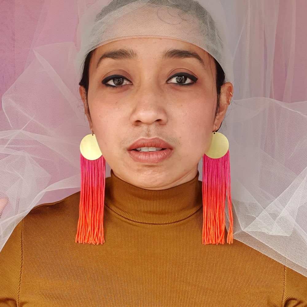 A model wears elegant pink and orange ombre tassel earrings with gold-plated round pendants. The bold, vibrant tassels add a statement touch, complementing the model's look with a stylish and sophisticated flair