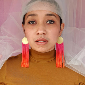 A model wears elegant pink and orange ombre tassel earrings with gold-plated round pendants. The bold, vibrant tassels add a statement touch, complementing the model's look with a stylish and sophisticated flair