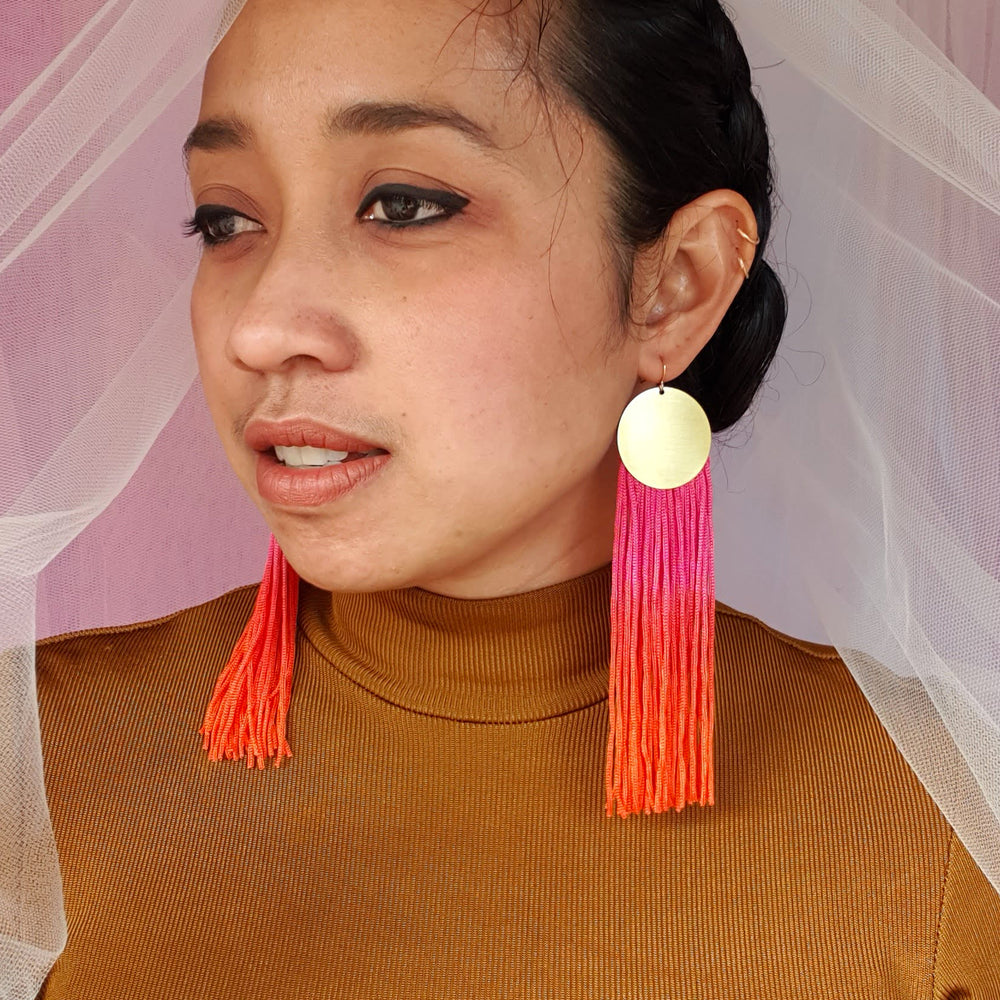 A model wears elegant pink and orange ombre tassel earrings with gold-plated round pendants. The bold, vibrant tassels add a statement touch, complementing the model's look with a stylish and sophisticated flair