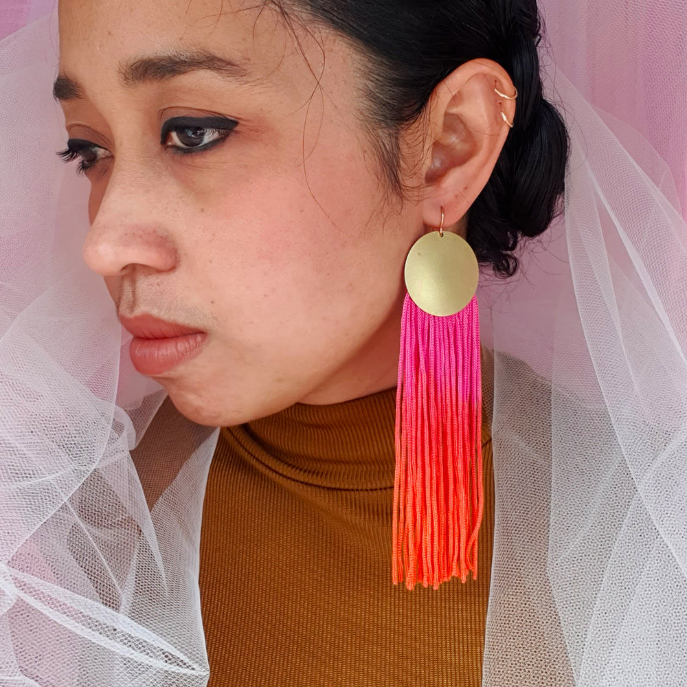 A model wears elegant pink and orange ombre tassel earrings with gold-plated round pendants. The bold, vibrant tassels add a statement touch, complementing the model's look with a stylish and sophisticated flair